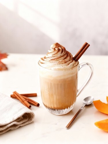 Eine Tasse Pumpkin Spice Latte mit Milchschaum, Zimt und Kürbisstücken auf einem herbstlich gedeckten Tisch.